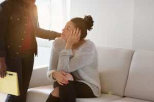 woman standing comforting another woman sitting on a couch talking about the warning signs for self-harm